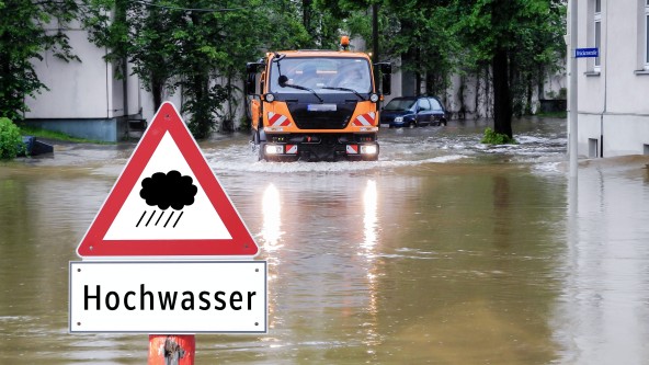 Ein orangener LKW fährt auf einer überfluteten Straße. Im Vordergrund ist ein Warnschild für Hochwasser zu sehen.